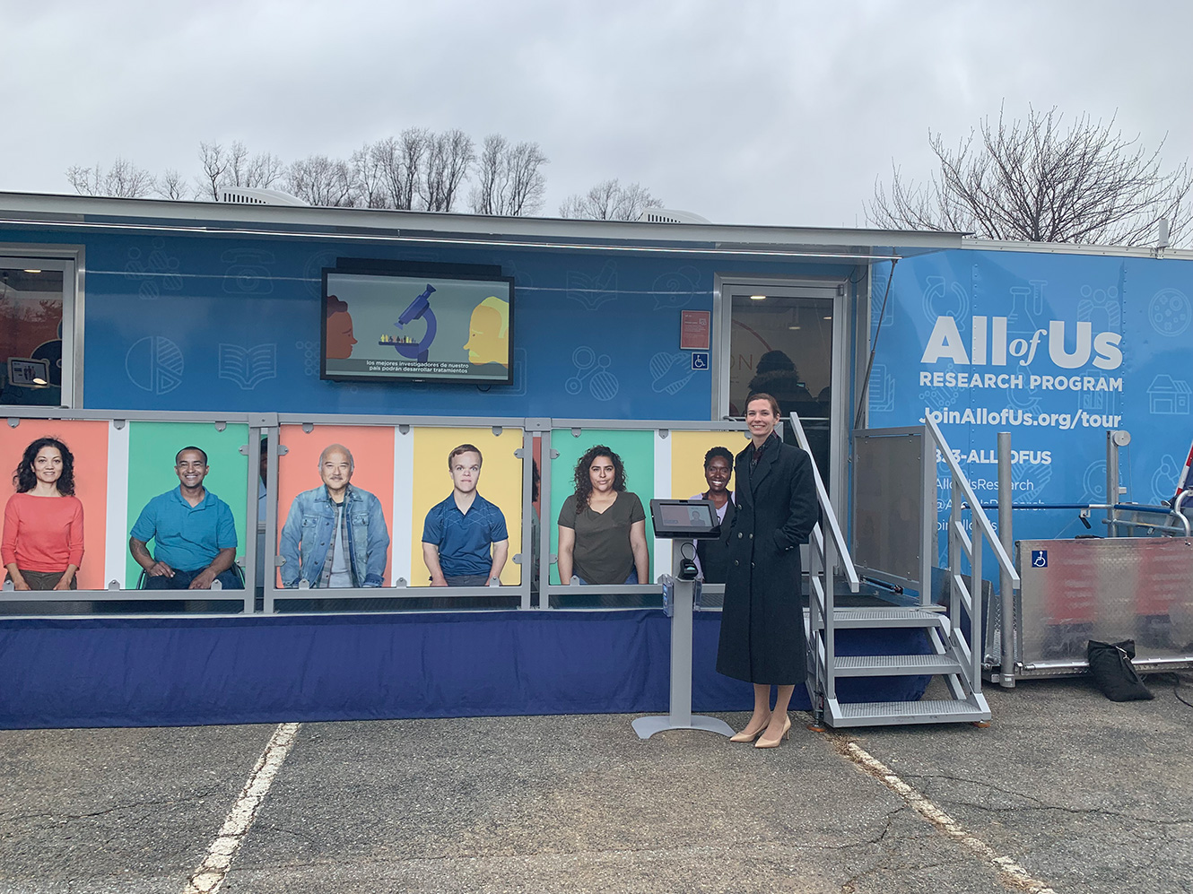 Adele is smiling as she stands outside a trailer that is covered in posters featuring many different people. On the trailer, a sign reads, All of Us Research Program.
